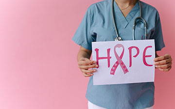 Healthcare worker in blue scrubs and a stethoscope holding a handwritten HOPE sign, with a pink awareness ribbon forming the letter O